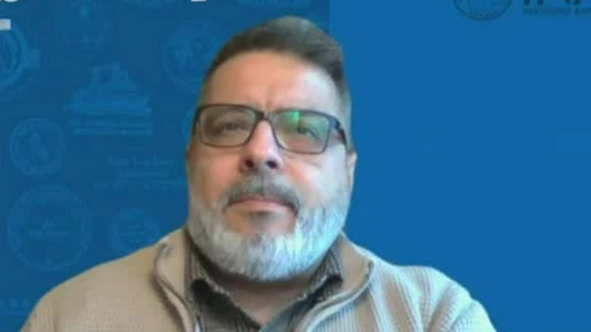 Hombre de mediana edad con anteojos y barba canosa durante una entrevista en canal de noticias, fondo azul con gráficos.