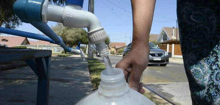 Una persona llena un bidón plástico con agua desde un grifo público instalado en la calle de un barrio residencial.