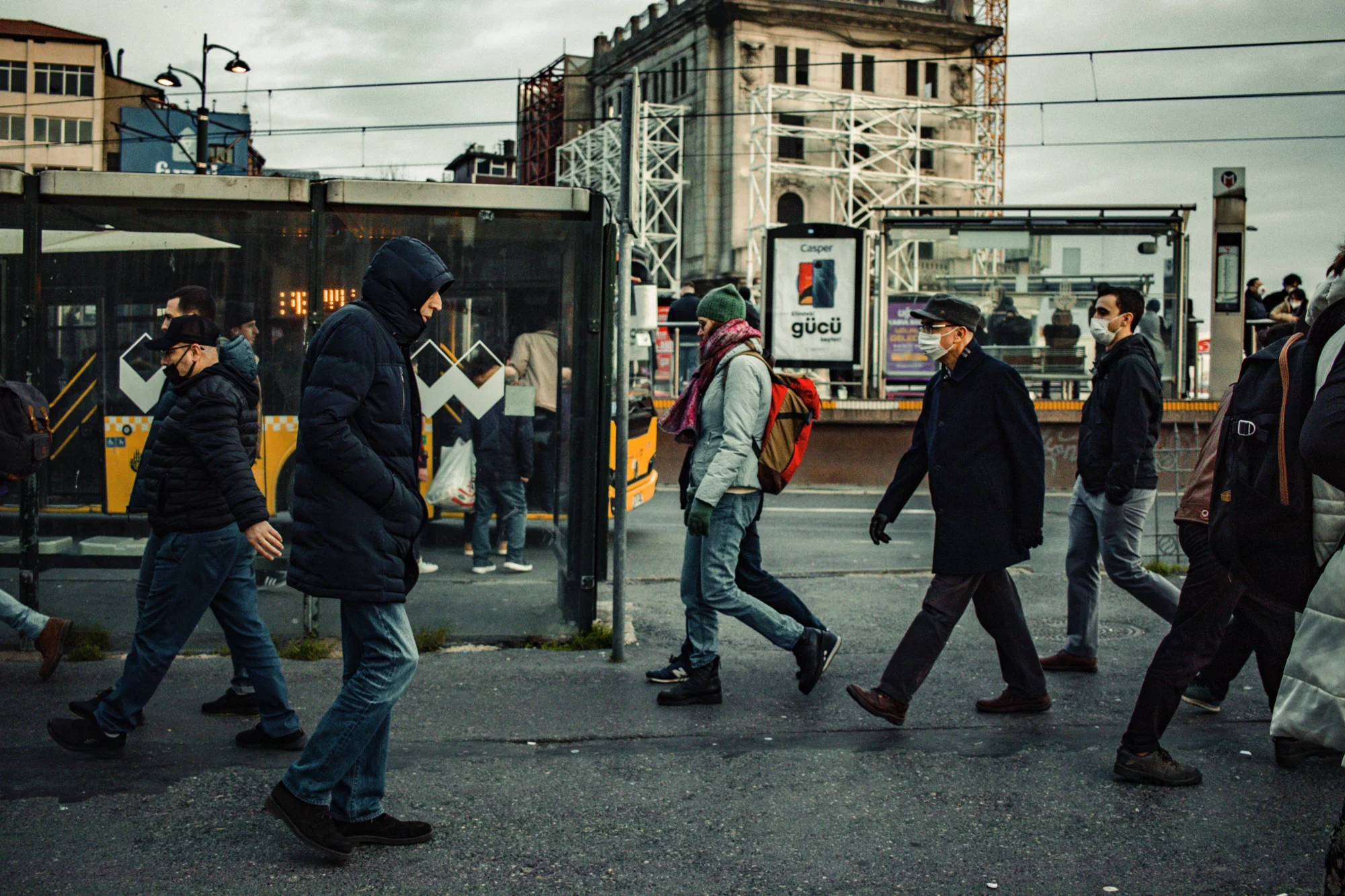 Grupo de personas con mascarilla y ropa de invierno cruza una calle concurrida junto a una parada de autobús en una ciudad turca.