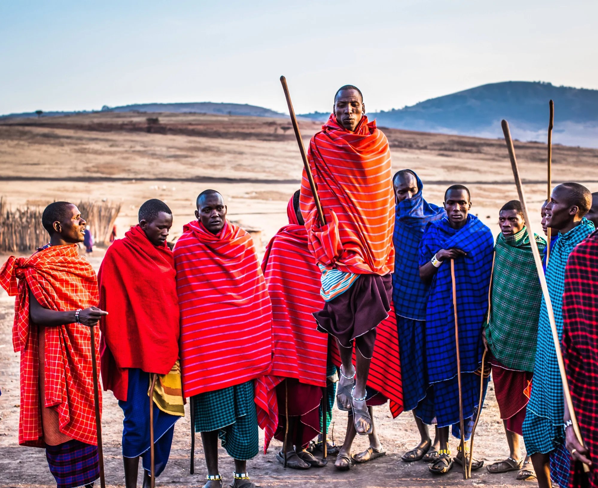 Grupo de guerreros masái con mantas de colores vivos y bastones; uno salta en el centro durante una ceremonia tradicional en paisaje árido africano.