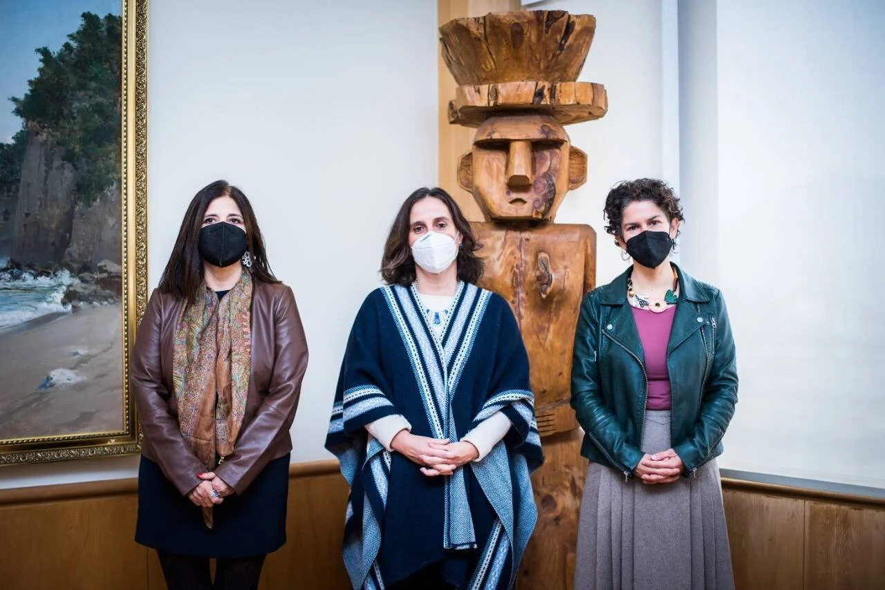 Tres mujeres con mascarilla posan frente a una escultura de madera tallada y un cuadro de paisaje costero.