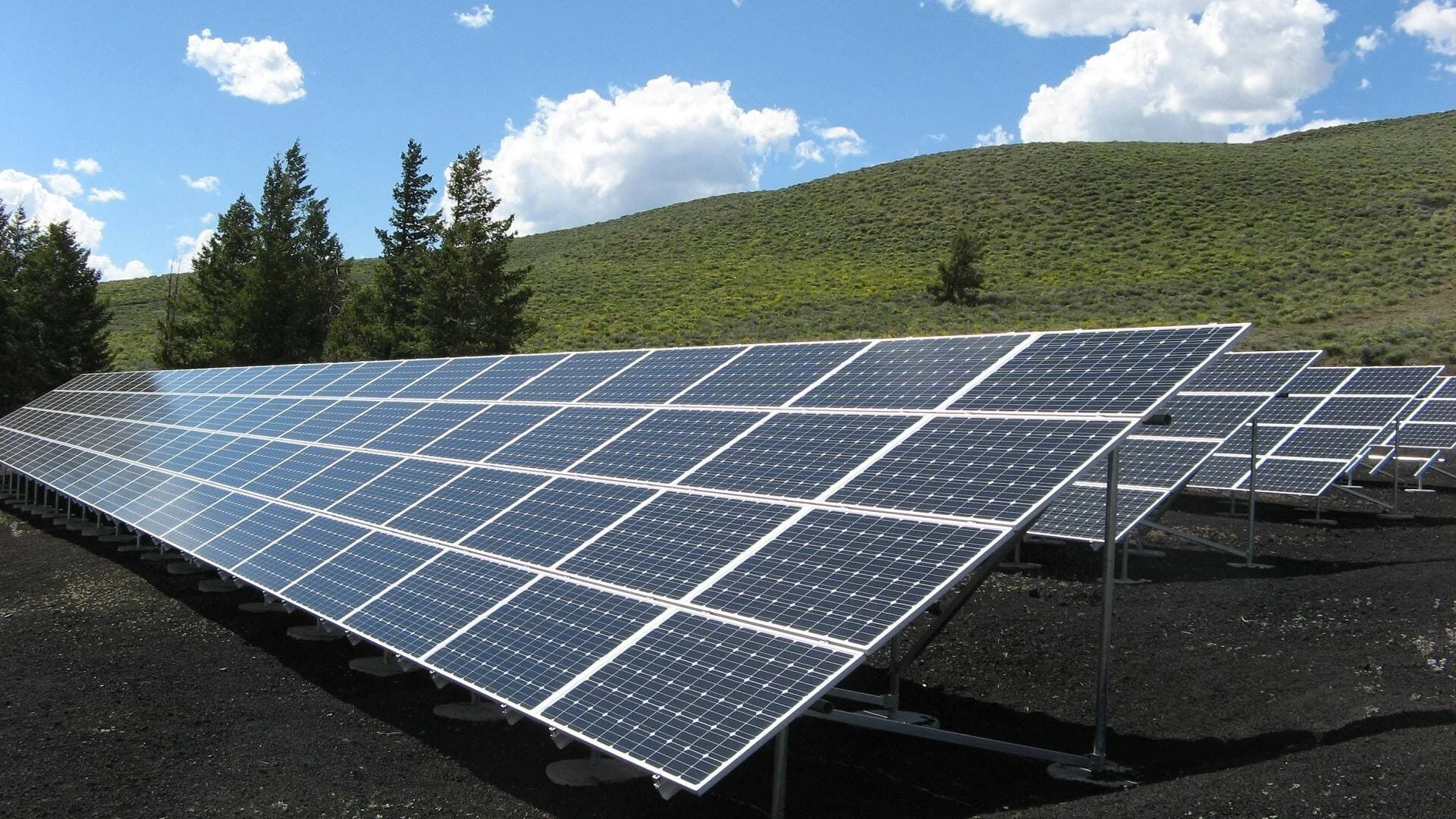 Filas de paneles solares fotovoltaicos instalados en el suelo frente a una colina verde bajo cielo azul con nubes.
