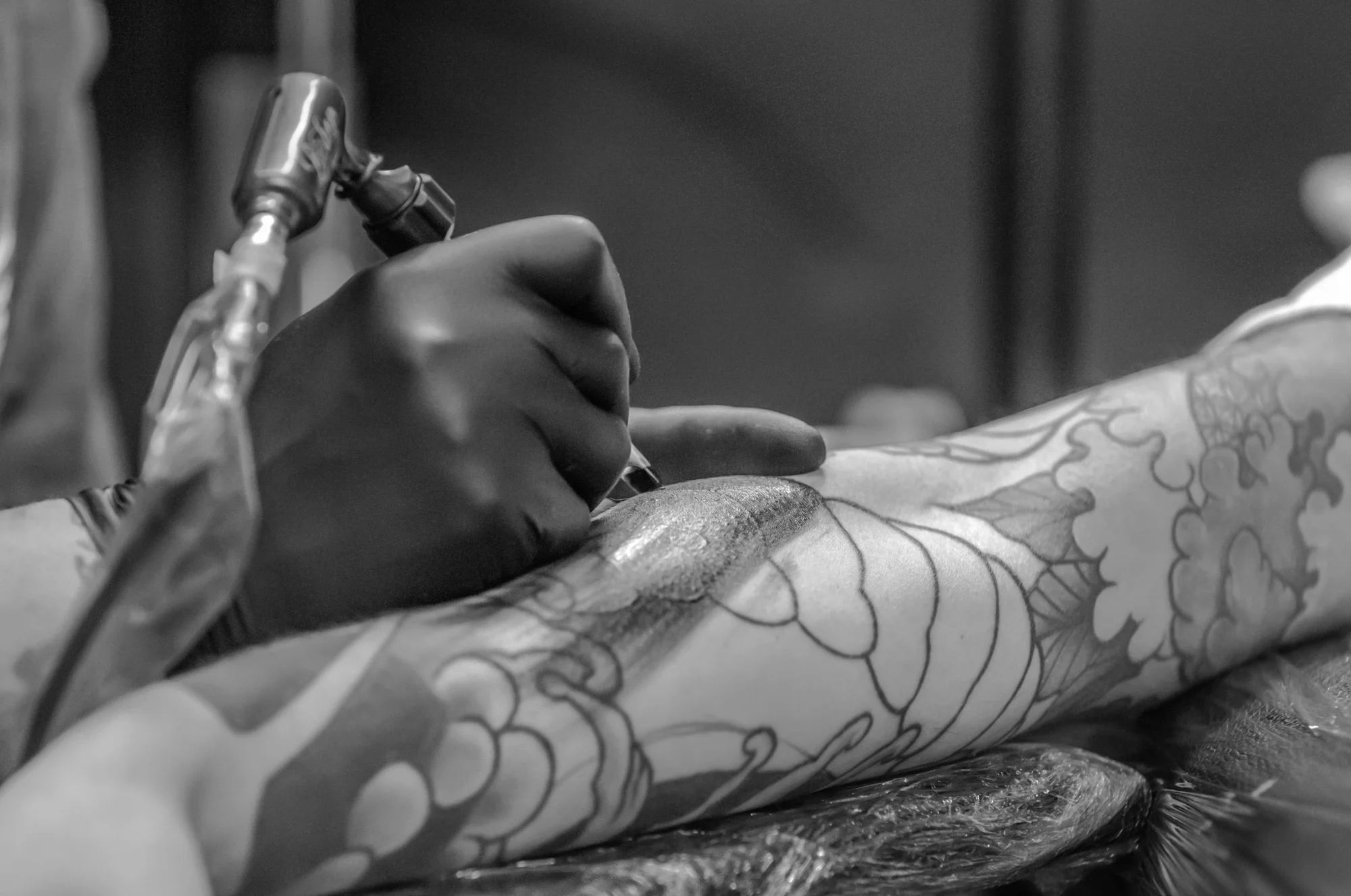 Tatuador con guantes negros usando máquina de tatuar sobre un brazo cubierto de diseños florales en proceso.