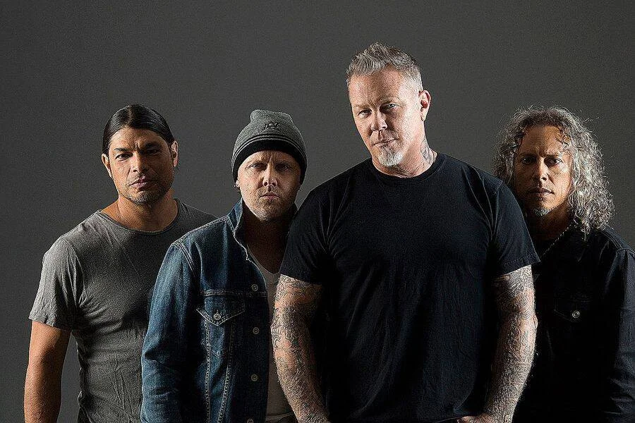Cuatro hombres adultos con expresión seria posan juntos frente a un fondo gris oscuro en una fotografía de banda musical.