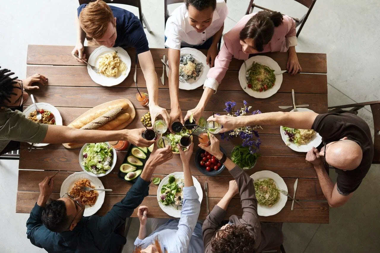 Vista cenital de un grupo de personas brindando con copas sobre una mesa llena de platos con ensaladas, pasta y pan.