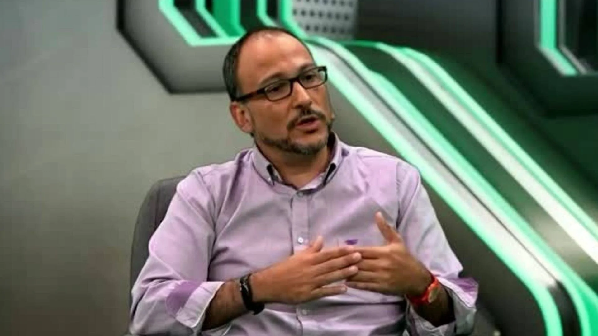 Hombre con lentes y camisa lila gesticula mientras habla sentado en un estudio de televisión con fondo verde.