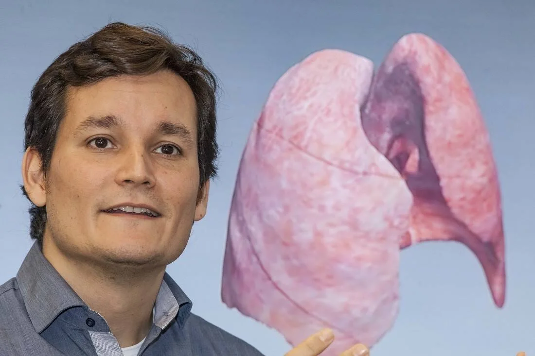 Un hombre sonríe frente a un modelo o imagen tridimensional de pulmones humanos de color rosado.