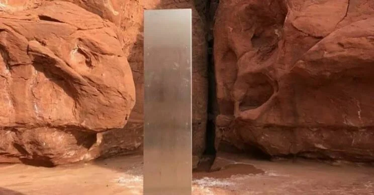 Columna metálica plateada y brillante erguida en medio de un cañón de rocas rojas en el desierto.