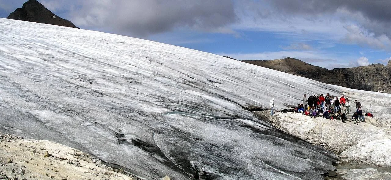 Un grupo de personas se detiene al borde de un extenso glaciar de alta montaña bajo un cielo parcialmente nublado.