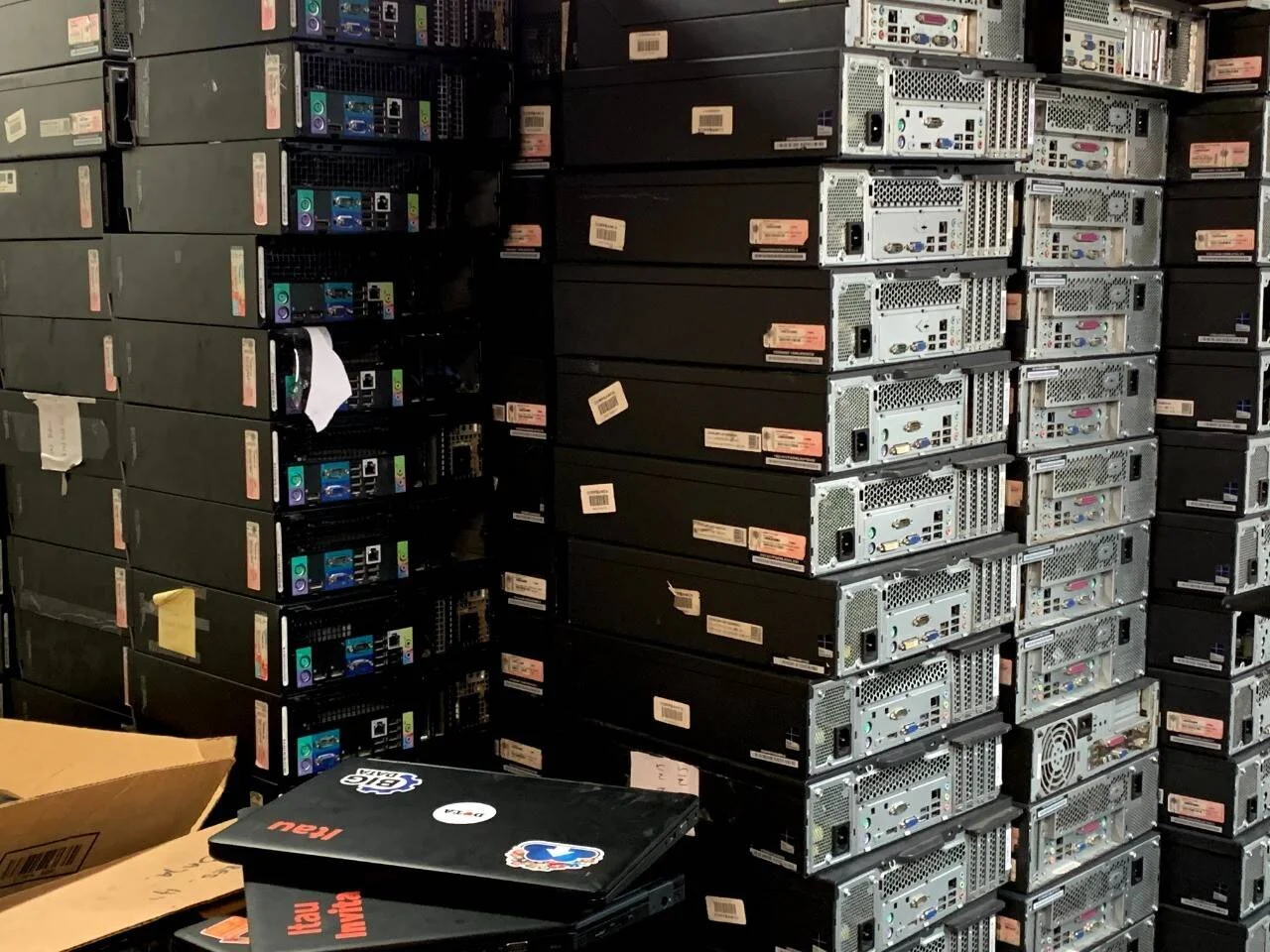 Decenas de gabinetes de computadoras de escritorio apilados hasta el techo en un depósito, con etiquetas de inventario visibles en sus paneles traseros.