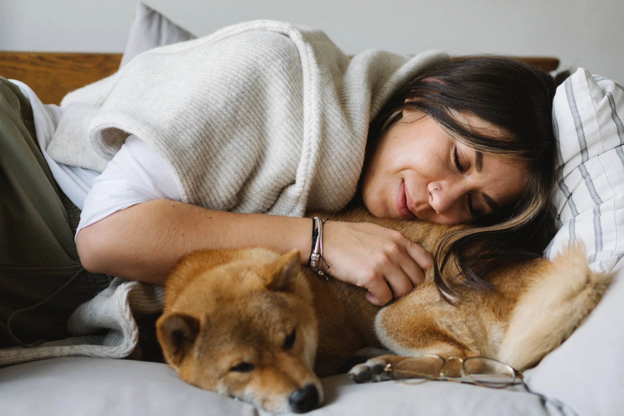 Mujer sonriente recostada en la cama abraza con ternura a un perro Shiba Inu mientras ambos descansan.