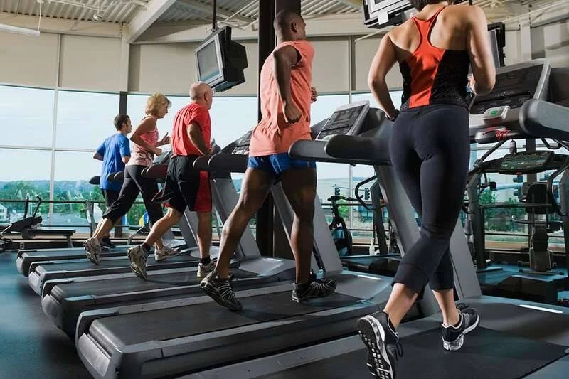Grupo de personas de distintas edades corriendo en cintas de correr en un gimnasio amplio e iluminado.
