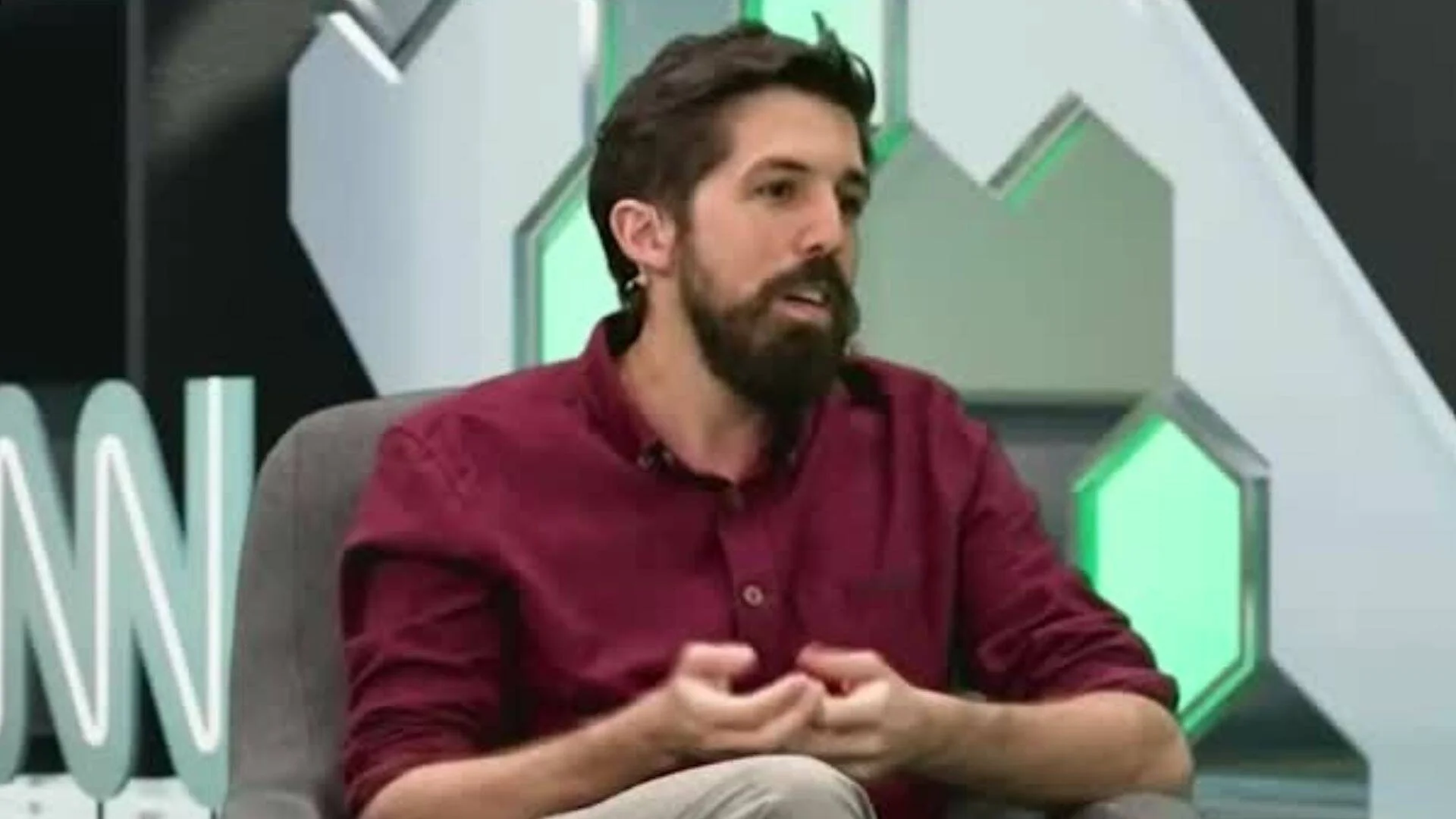 Hombre joven con barba y camisa bordó habla sentado en un estudio de televisión con decorado verde y gris.