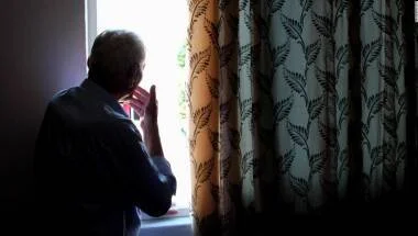 Persona mayor de espaldas observa por una ventana junto a una cortina con estampado de hojas.