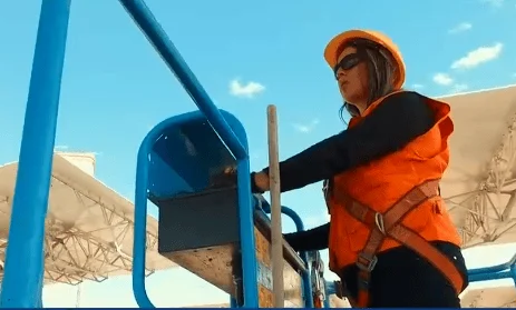 Mujer con casco naranja, lentes y arnés de seguridad sube una escalera metálica azul en obra.