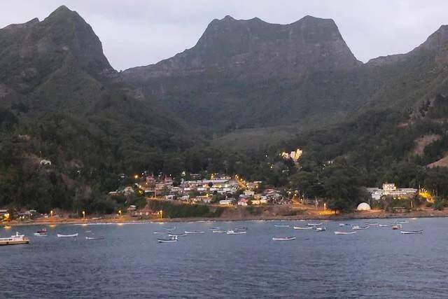 Pequeño pueblo costero con luces encendidas al atardecer, rodeado de montañas, con botes anclados en la bahía.