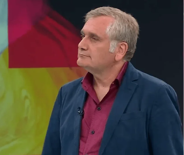 Hombre de cabello canoso con saco azul y camisa burdeos de pie en un estudio de televisión con fondo colorido.