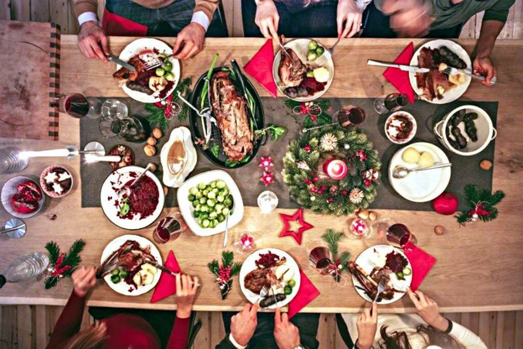 Vista cenital de una mesa navideña con platos de carne, coles de Bruselas, vino tinto y decoración festiva mientras varias personas comen.