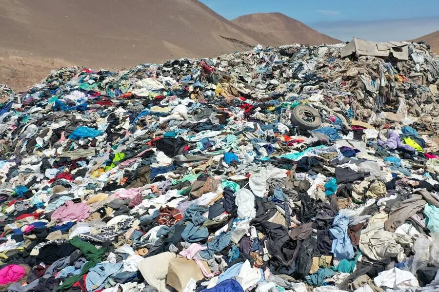 Montaña de ropa descartada acumulada en un desierto, con cerros áridos al fondo, evidenciando la contaminación de la industria textil.