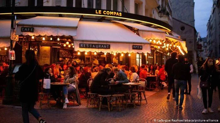 Personas sentadas en la terraza exterior del café-restaurante Le Compas de París durante la noche, con luces cálidas y ambiente animado.