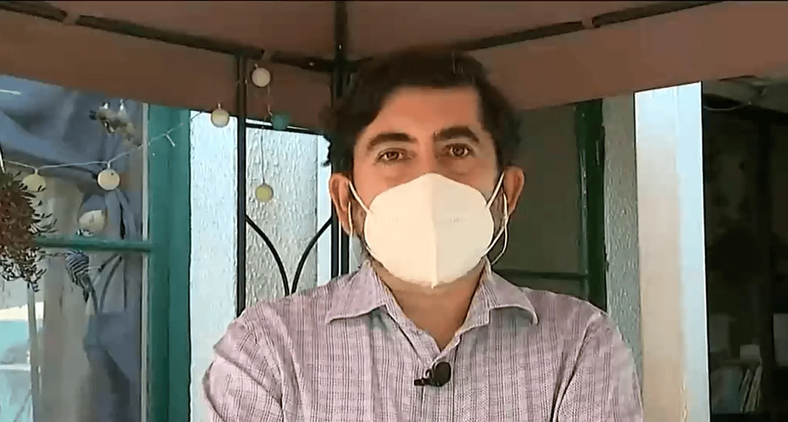 Hombre con mascarilla blanca KN95 y micrófono de solapa habla frente a cámara desde un espacio exterior techado.
