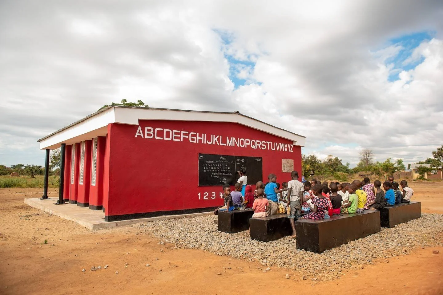 Grupo de niños recibe clase al aire libre frente a una escuela rural de pared roja con el abecedario pintado.