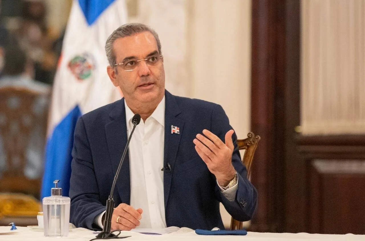 Hombre con traje azul y pin de la bandera dominicana habla ante un micrófono frente a la bandera de República Dominicana.