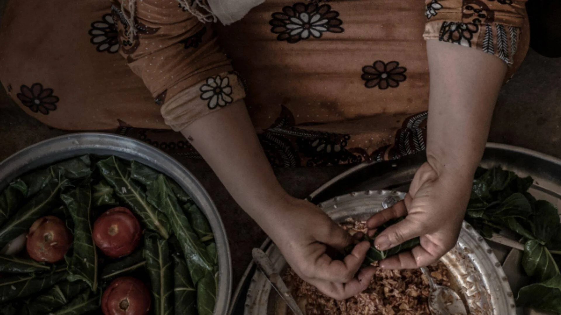 Manos de una mujer envolviendo relleno en hojas verdes, junto a fuentes metálicas con frutas rojas y vegetales.