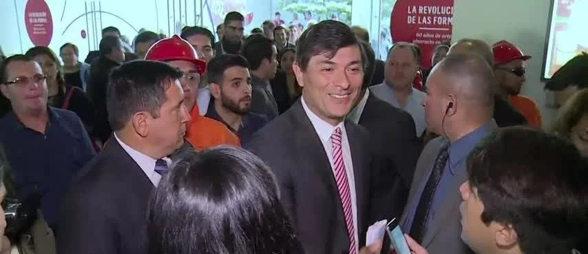 Un hombre con traje y corbata a rayas sonríe rodeado de periodistas y trabajadores con cascos rojos en un evento institucional.