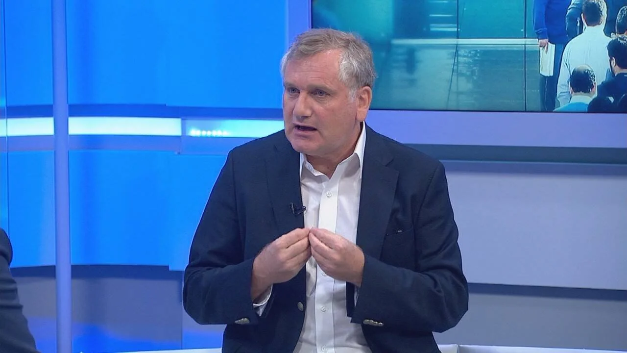 Hombre de traje oscuro y camisa blanca habla gesticulando en un estudio de televisión con fondo azul.