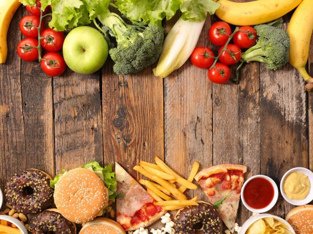 Mesa de madera con frutas y verduras frescas en la parte superior y hamburguesa, pizza, papas fritas y donas en la inferior, ilustrando la elección alimentaria.