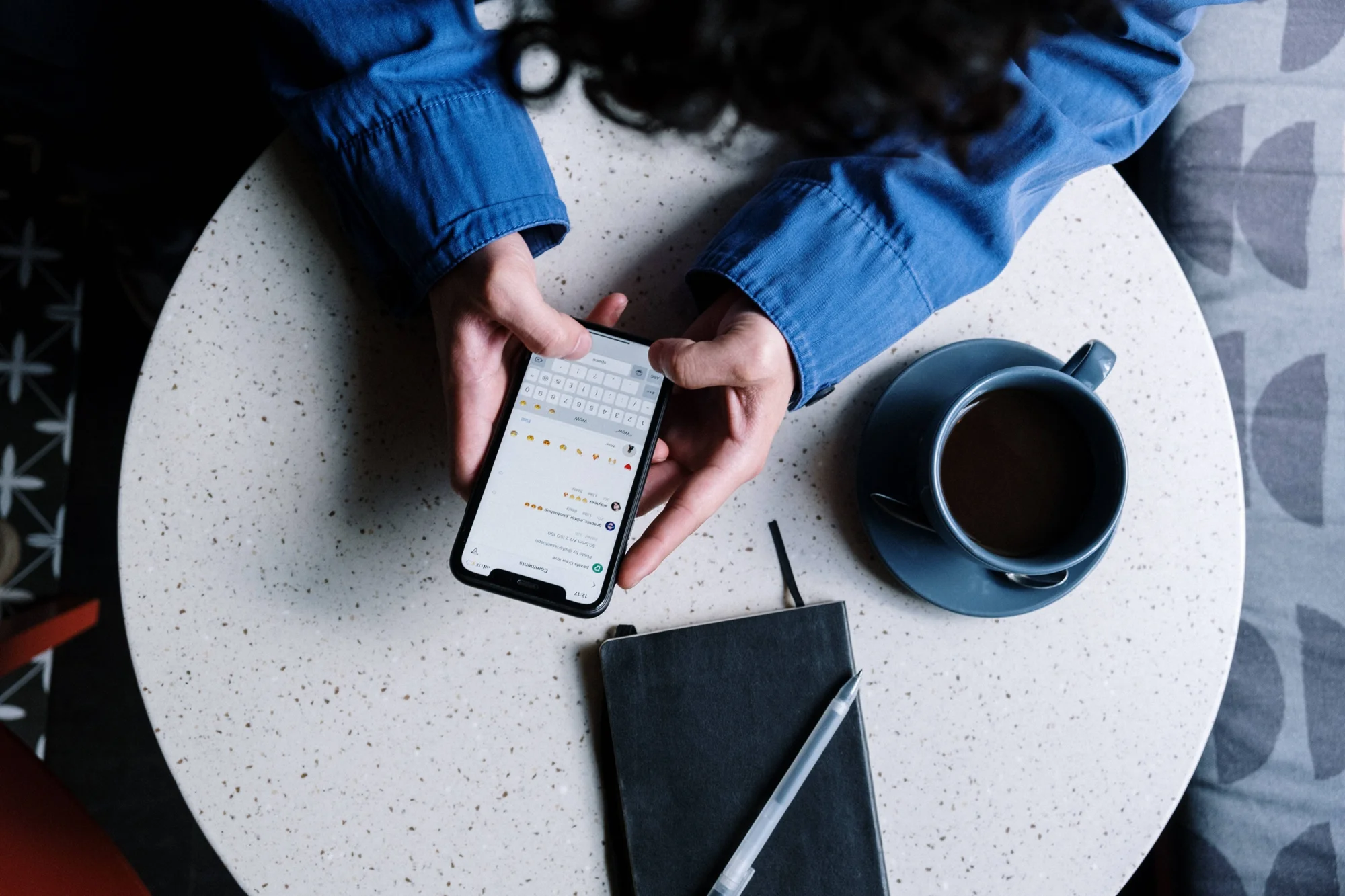 Vista cenital de una persona escribiendo en su teléfono con emojis visibles en pantalla, junto a una taza de café y una libreta sobre una mesa redonda.