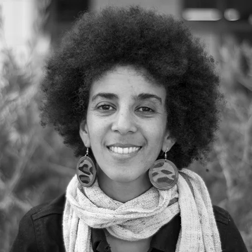 Mujer joven con cabello afro, aretes redondos y bufanda, sonriendo en exterior, fotografía en blanco y negro.