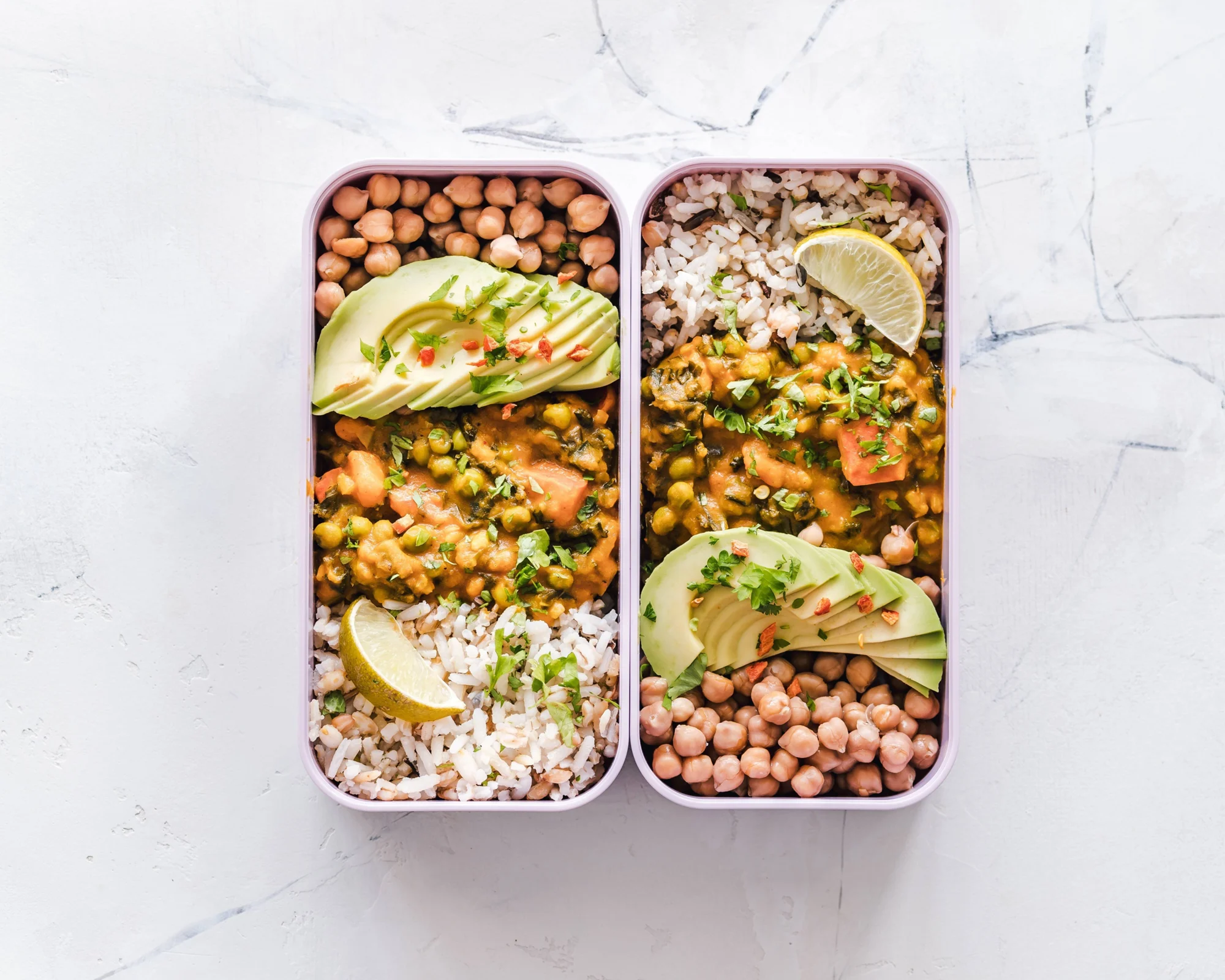 Dos recipientes con curry de garbanzos, arroz integral, aguacate en láminas, cilantro y rodajas de limón, vistos desde arriba.