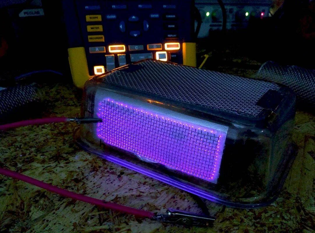 Dispositivo rectangular con rejilla metálica y matriz de LEDs violetas encendidos, conectado con cables a un equipo de medición electrónico.