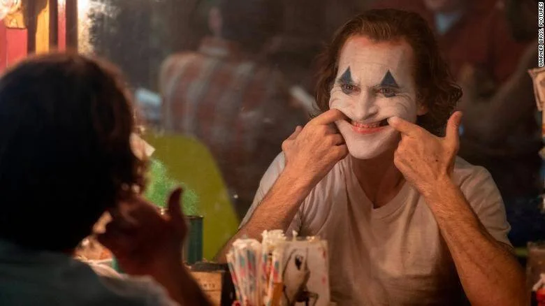 Actor con maquillaje blanco y sonrisa pintada del Joker se estira las comisuras de la boca frente a otro personaje en una escena de la película.