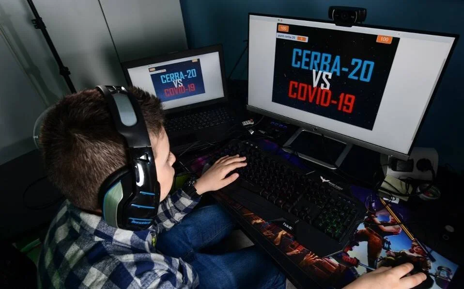 Un niño con auriculares gaming juega en una computadora con pantallas que muestran el título 'CERRA-20 vs COVID-19'.