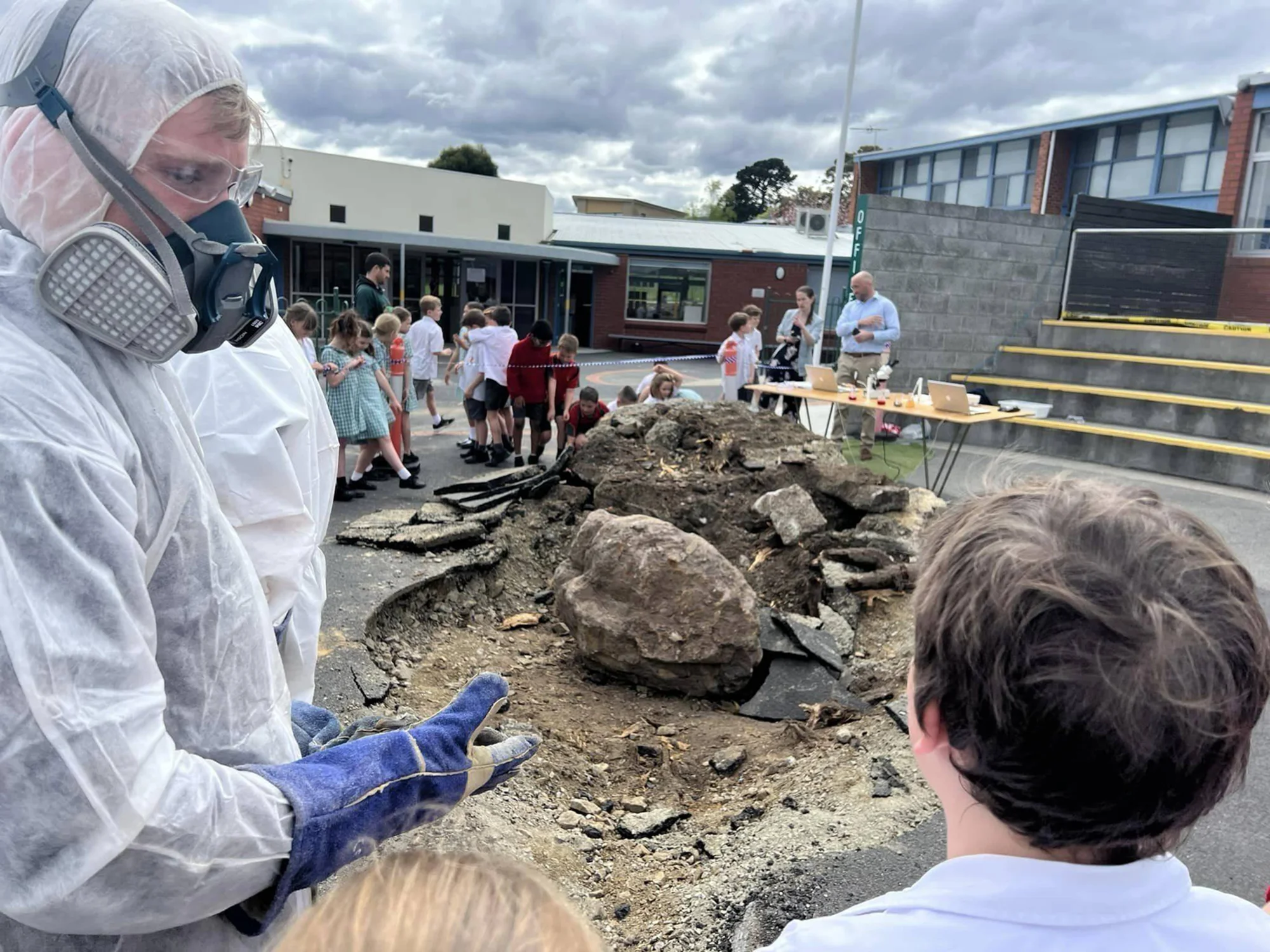 Un técnico con traje hazmat, mascarilla y guantes supervisa una excavación con rocas y tierra en el patio de una escuela rodeado de niños.