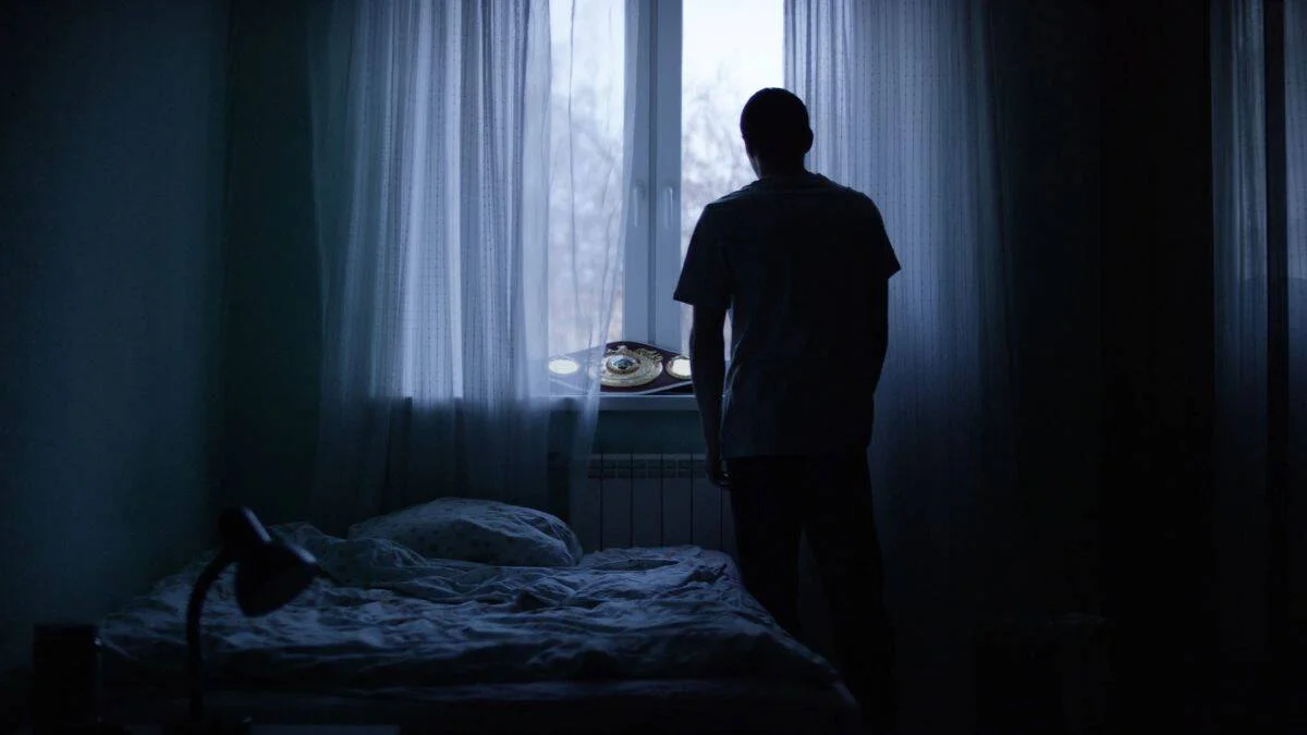 Silueta de un hombre de pie en habitación oscura mirando por la ventana, con un cinturón de campeón de boxeo apoyado en el alféizar.
