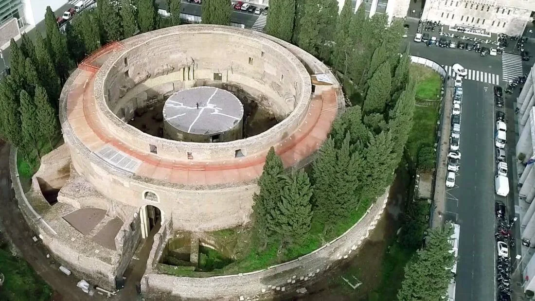 Vista aérea de un gran mausoleo romano circular de piedra rodeado de árboles y calles urbanas.