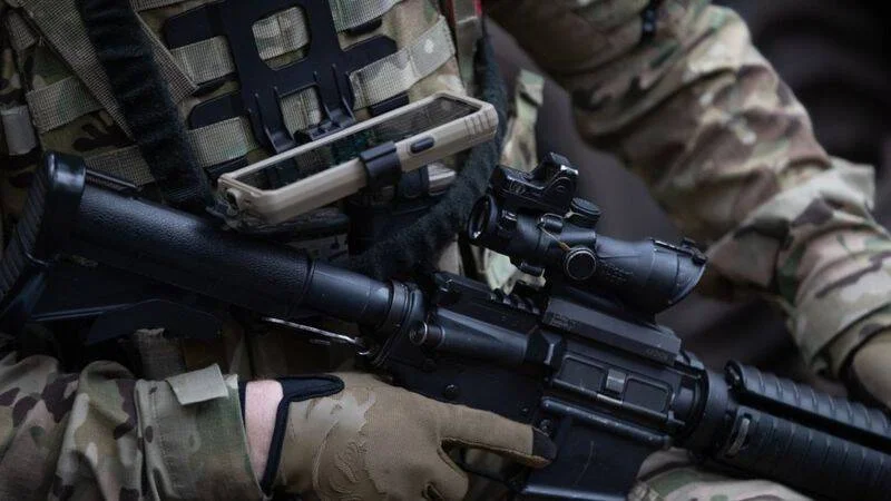 Dos militares con uniforme camuflado sostienen un rifle de asalto con mira óptica y portan equipo táctico.