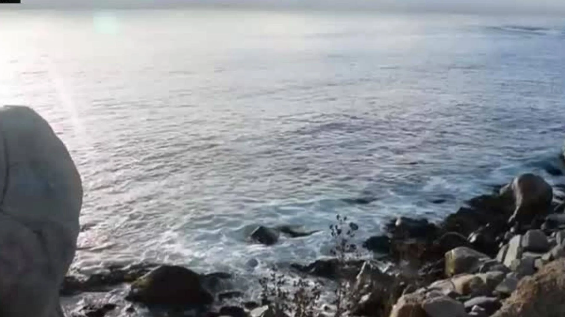 Orilla rocosa junto a un lago o mar en calma, con reflejo de luz solar sobre el agua en un día nublado.