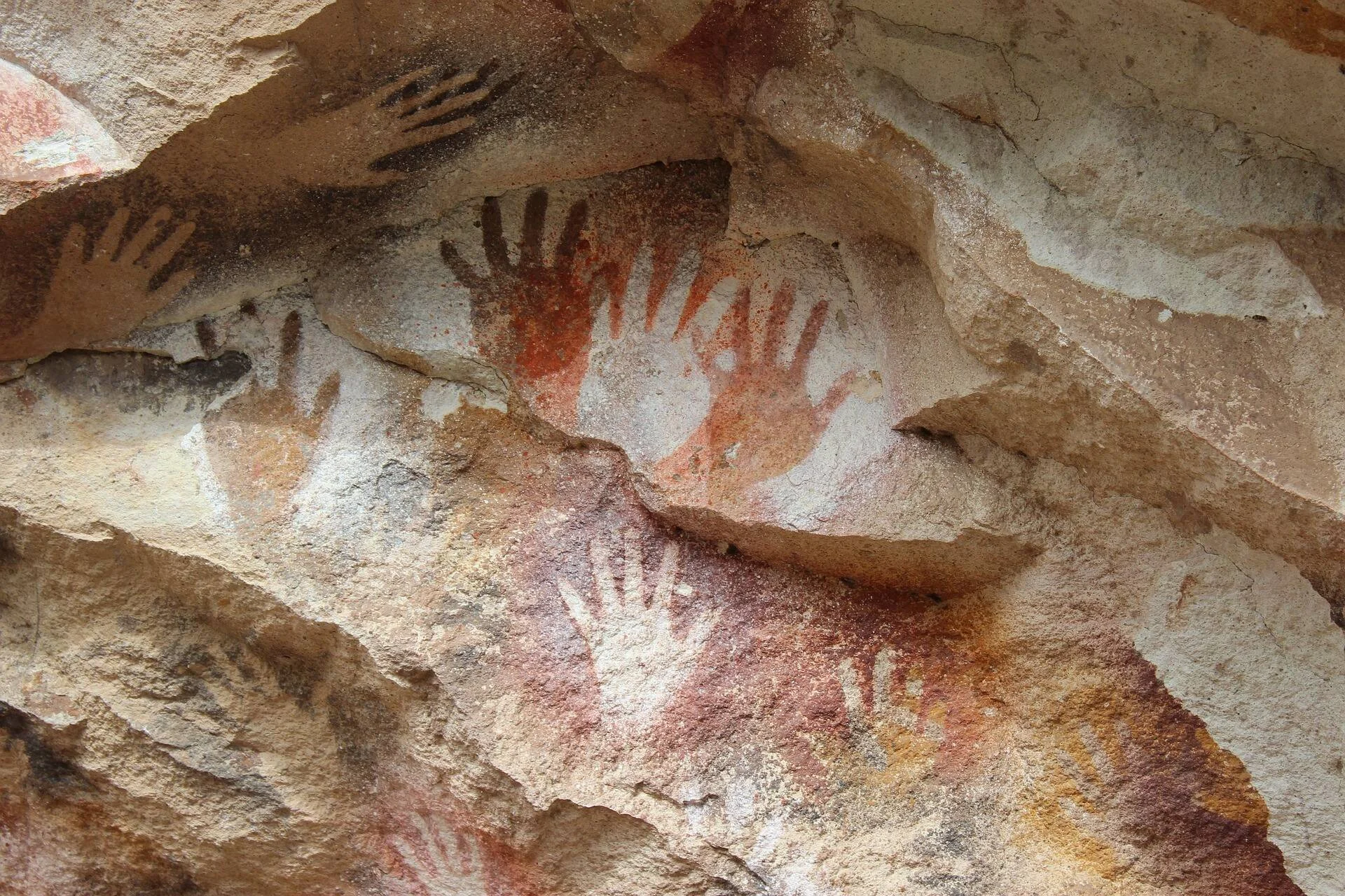 Múltiples impresiones de manos prehistóricas en colores rojo, blanco y negro estampadas sobre una pared de roca.