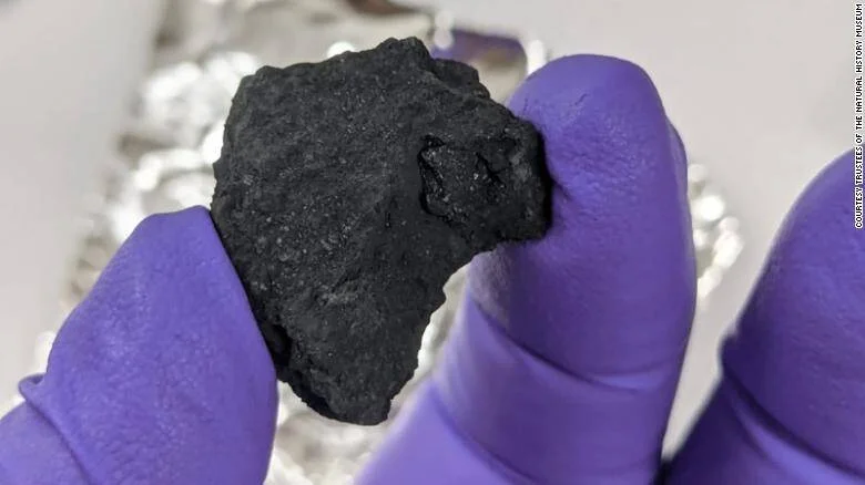 Manos con guantes morados sostienen un fragmento de meteorito negro y poroso sobre papel de aluminio.