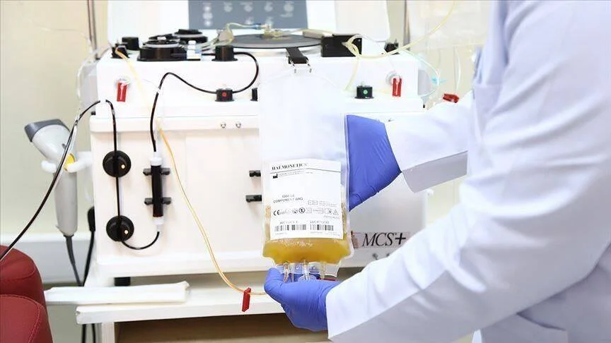 Un profesional sanitario con guantes sostiene una bolsa de plasma sanguíneo conectada a una máquina de aféresis en un laboratorio médico.