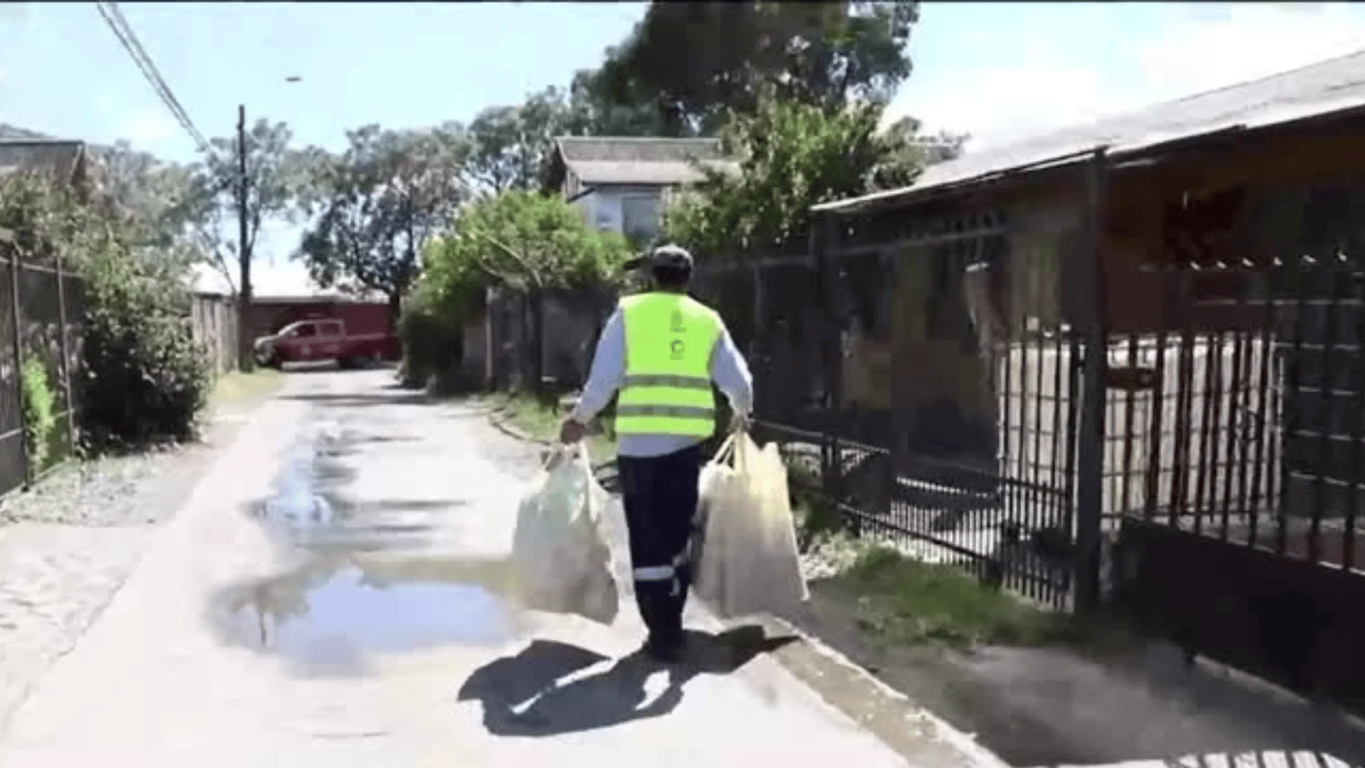 Trabajador con chaleco reflectante amarillo camina por un pasaje residencial llevando bolsas de ayuda a vecinos.