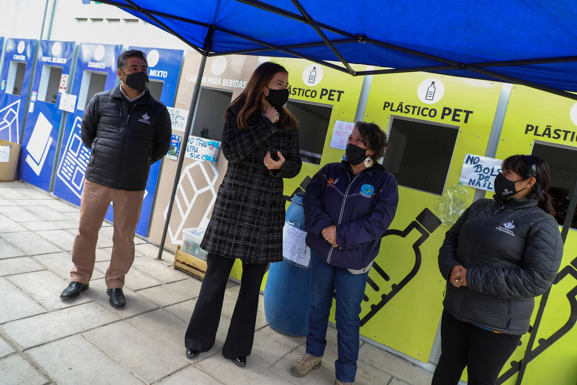Cuatro personas con mascarilla conversan frente a contenedores de reciclaje de plástico PET y cartón en un punto limpio municipal.
