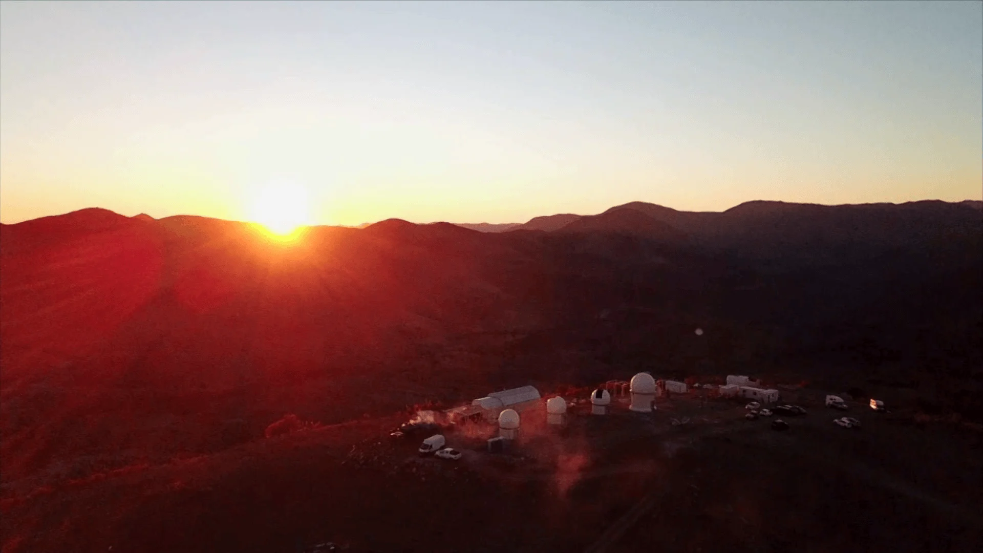 Vista aérea de un complejo de observatorio con cúpulas blancas en una cumbre montañosa durante la puesta de sol.