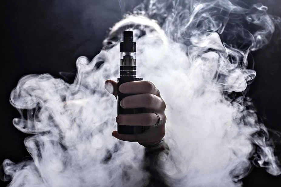 Una mano sostiene un dispositivo de vapeo rodeado de una densa nube de vapor blanco sobre fondo negro.