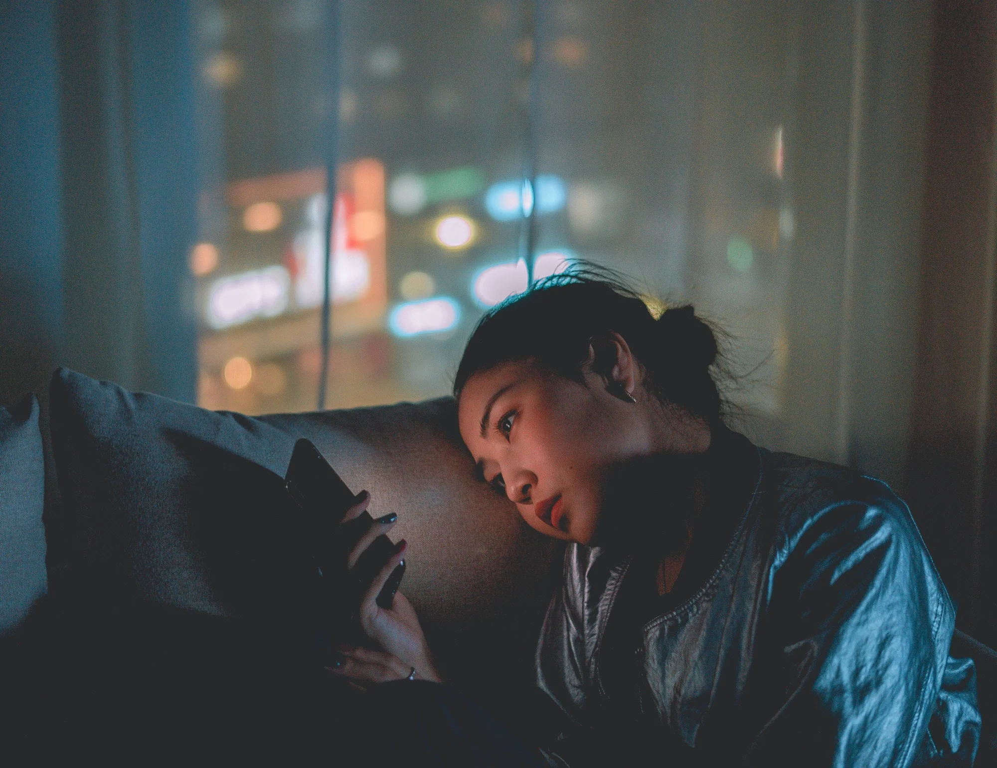 Mujer joven recostada en un sofá oscuro mira su teléfono con luces nocturnas de la ciudad al fondo.